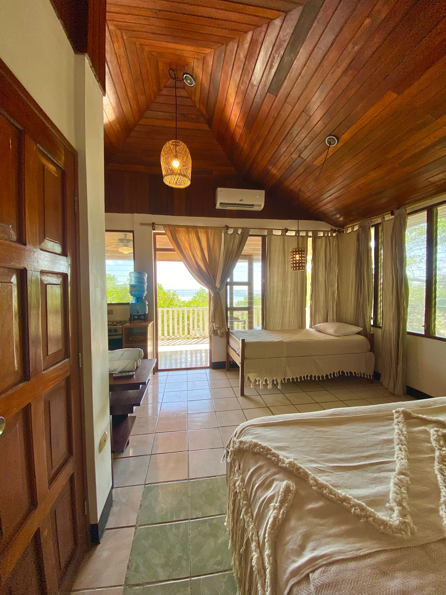 A room with two beds beneath a wood-panelled vaulted ceiling, opening onto a deck with an ocean view