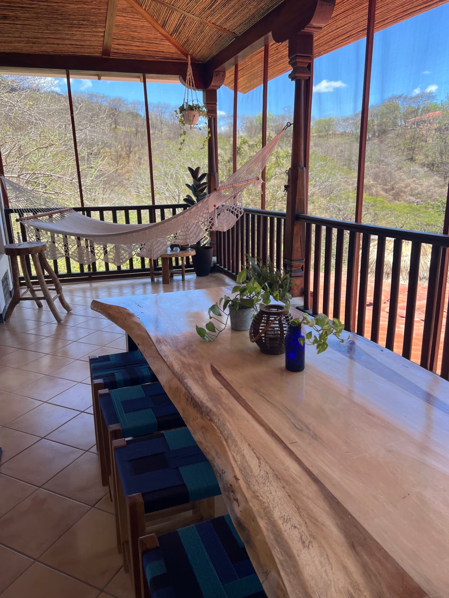 A long live-edge wooden table with woven stools beside a hammock on an open-air terrace