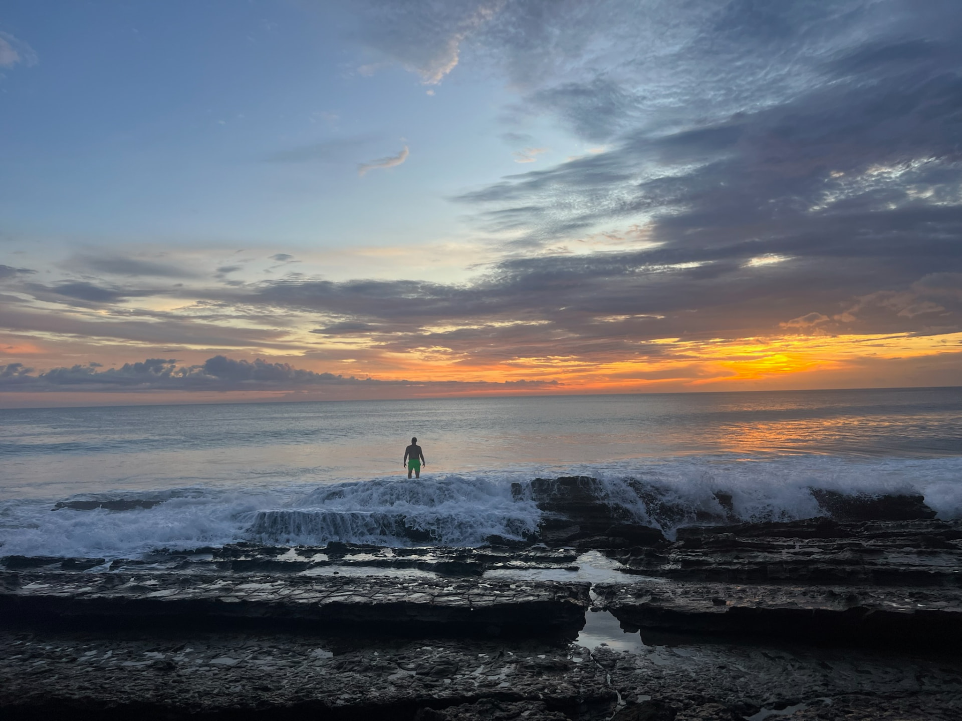 A person standing in the Pacific waves at sunset beside the rocky shoreline