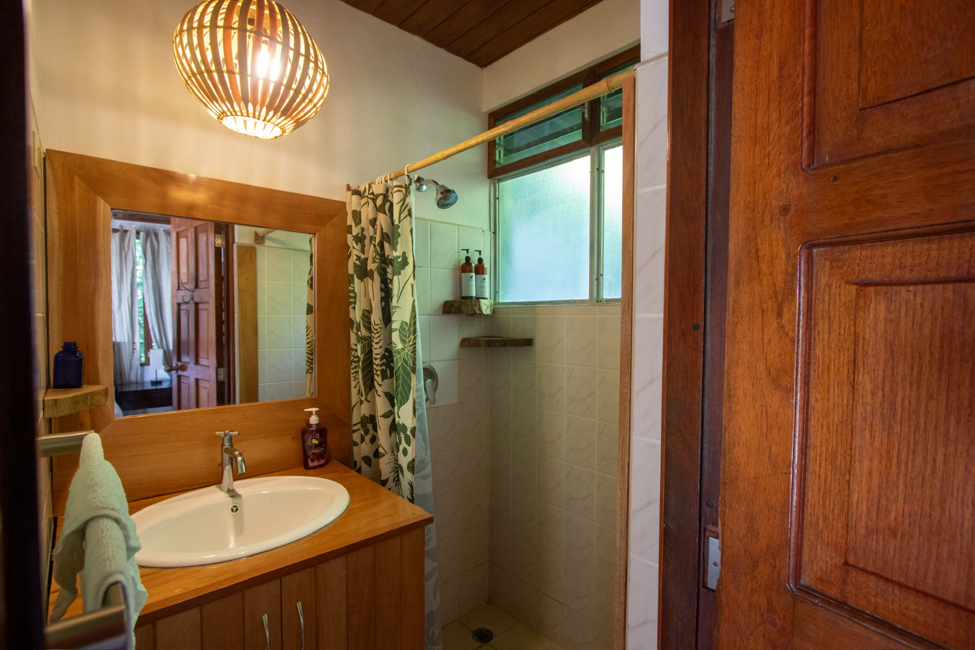 A tiled bathroom with a wooden vanity, mirror and shower beside a louvred window