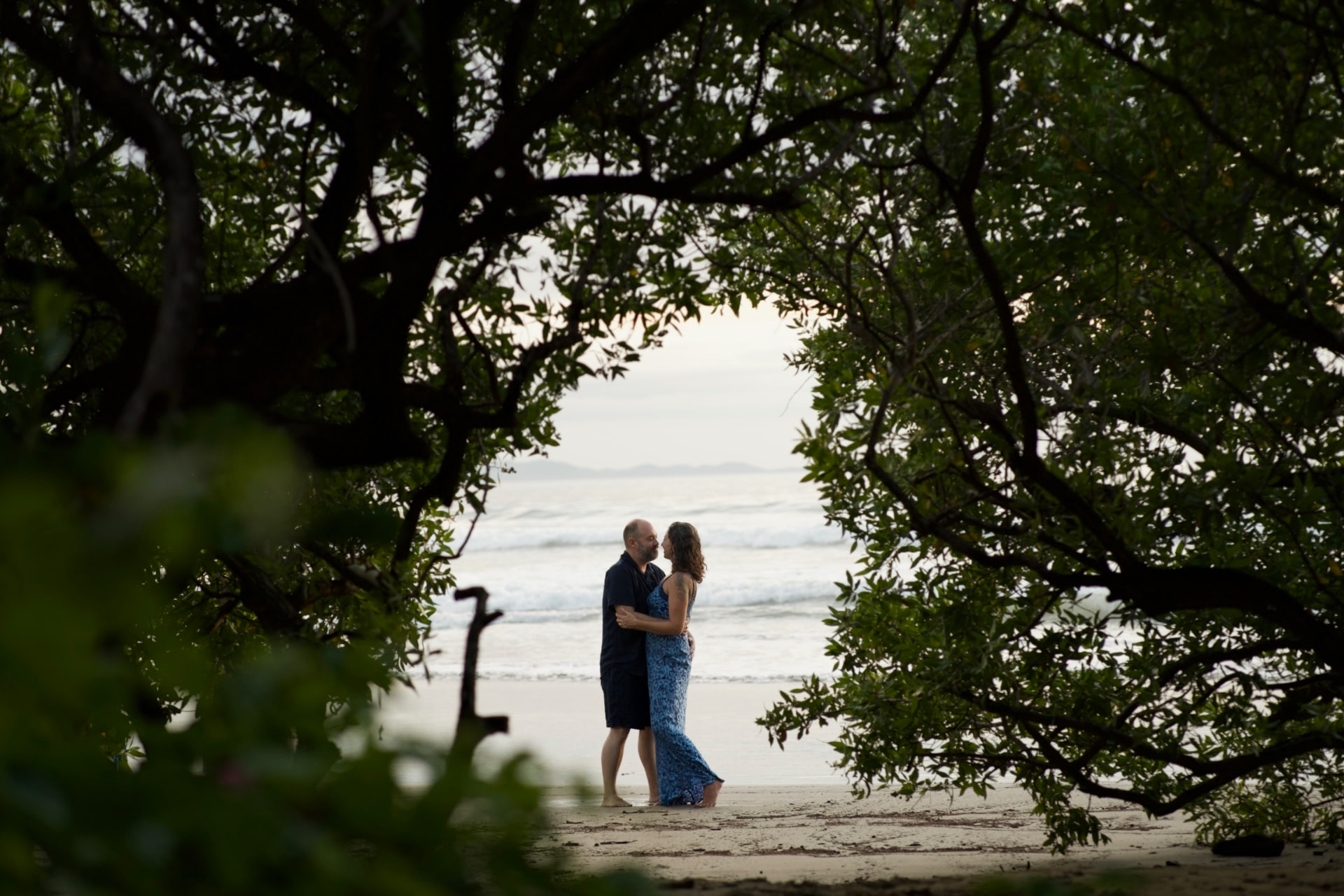A couple standing together on the beach, framed by dark leaves