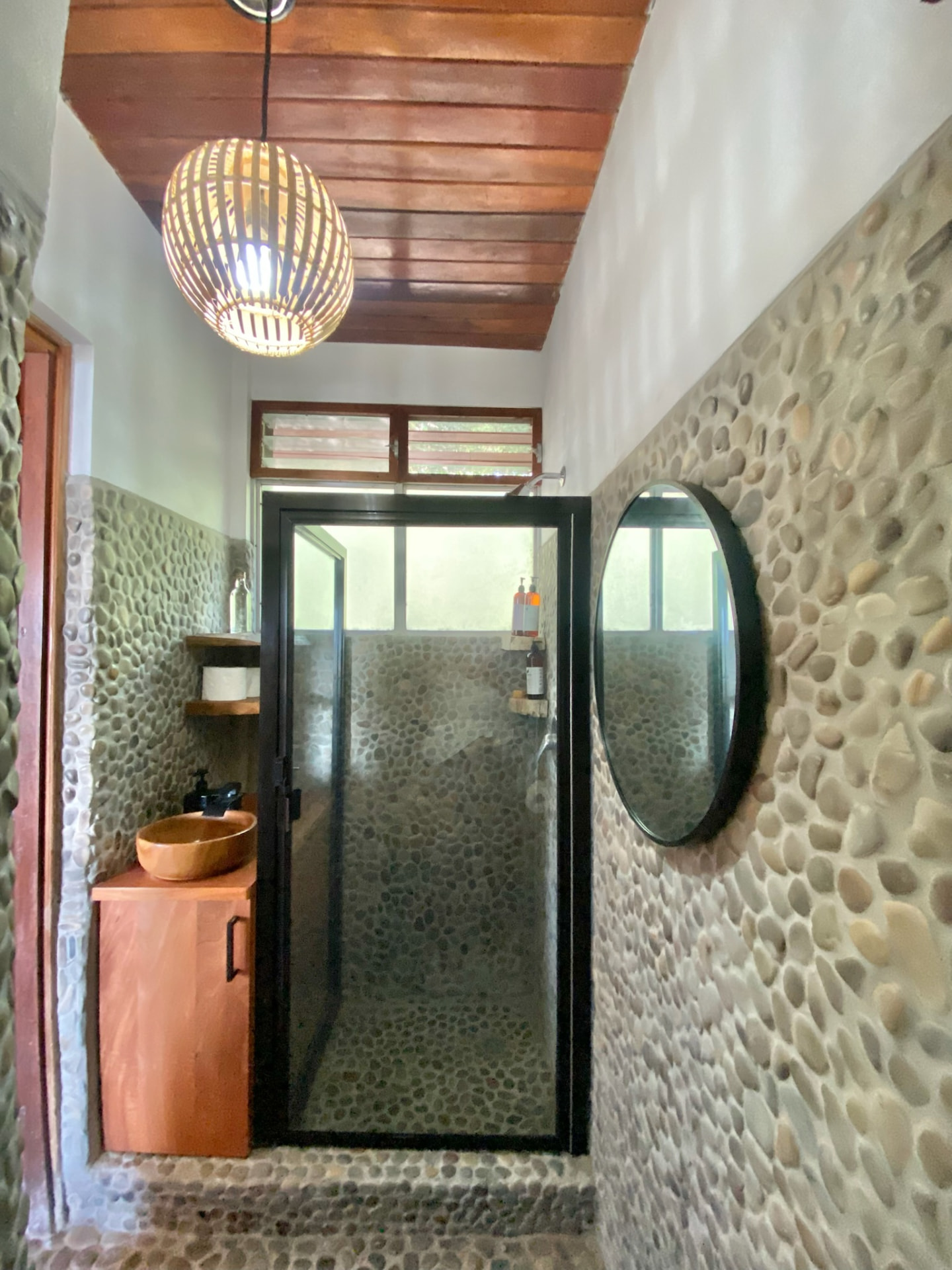 A bathroom with river-stone walls, a wooden vanity with a carved basin and a glass shower door
