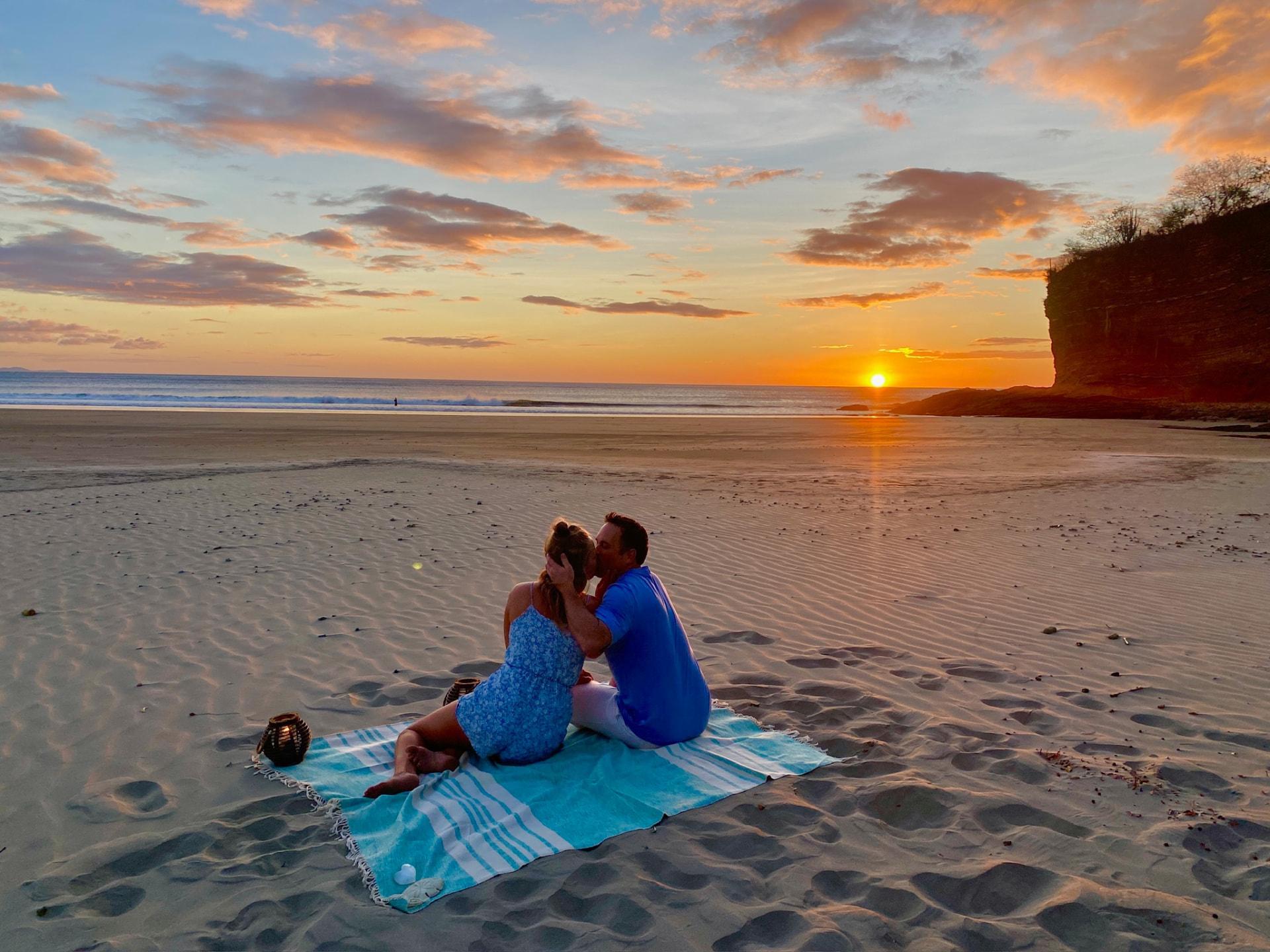 A couple embracing on a beach blanket as the sun sets beside the rocky shoreline